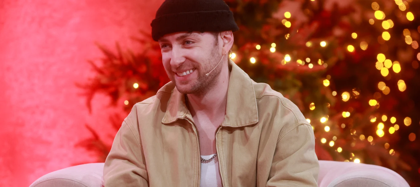 Dominik Dudek ogłosił radosną nowinę Uśmiechnięty mężczyzna w jasnej kurtce i czarnej czapce siedzi na jasnej sofie, w tle dekoracje świąteczne i choinka oświetlona ciepłym światłem.