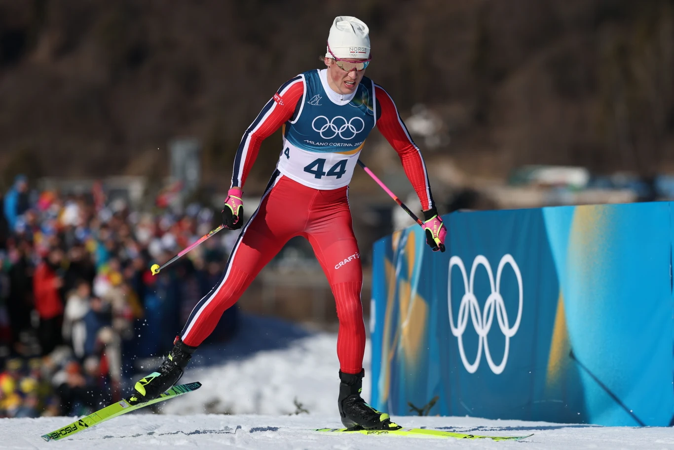 Zawodnik w czerwonym stroju narciarskim biegnący na nartach biegowych podczas zawodów olimpijskich, otoczony zimową scenerią, w tle widoczne są symbole olimpijskie i publiczność.