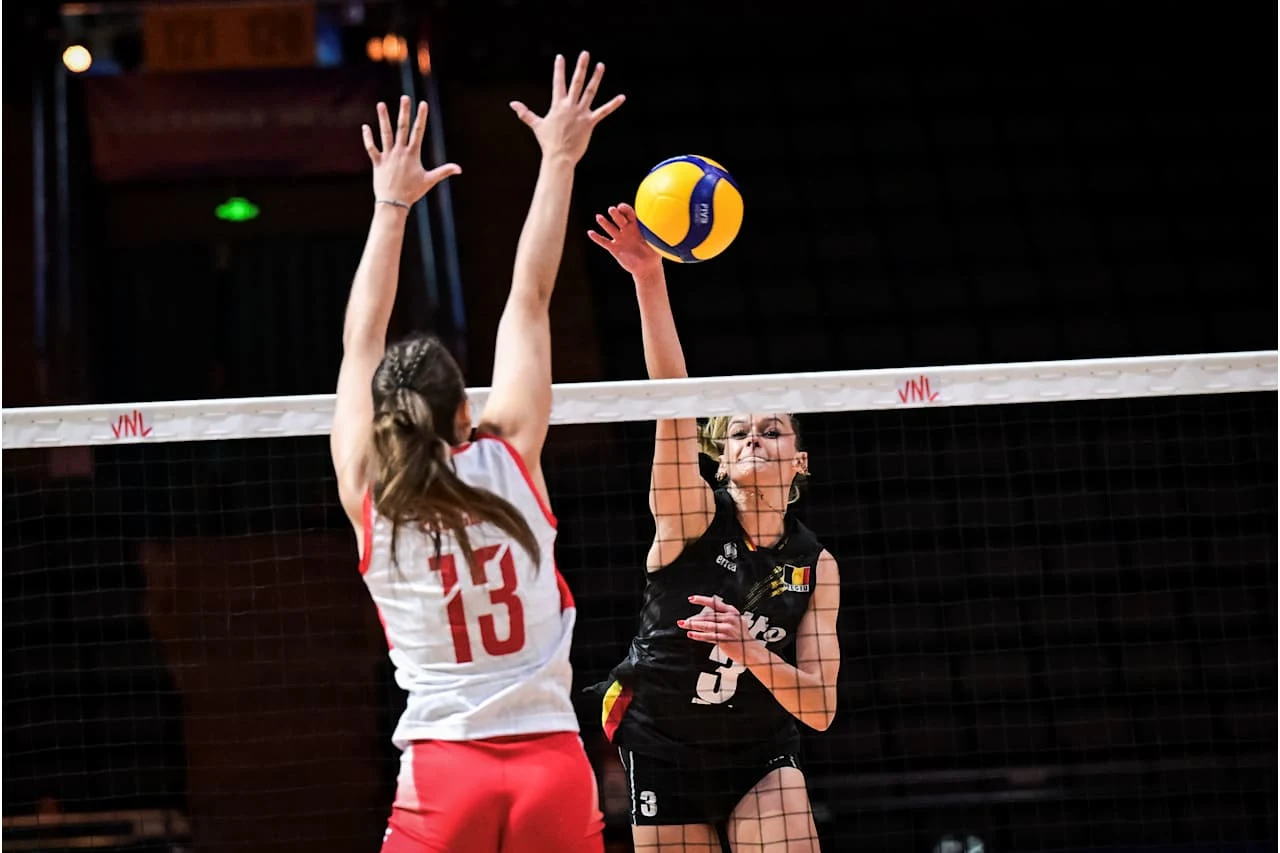 Zawodniczka w czarnym stroju wykonuje atak siatkarski przez siatkę, podczas gdy przeciwniczka w biało-czerwonym stroju próbuje ją zablokować, obie skoncentrowane na piłce unoszącej się tuż nad siatką.