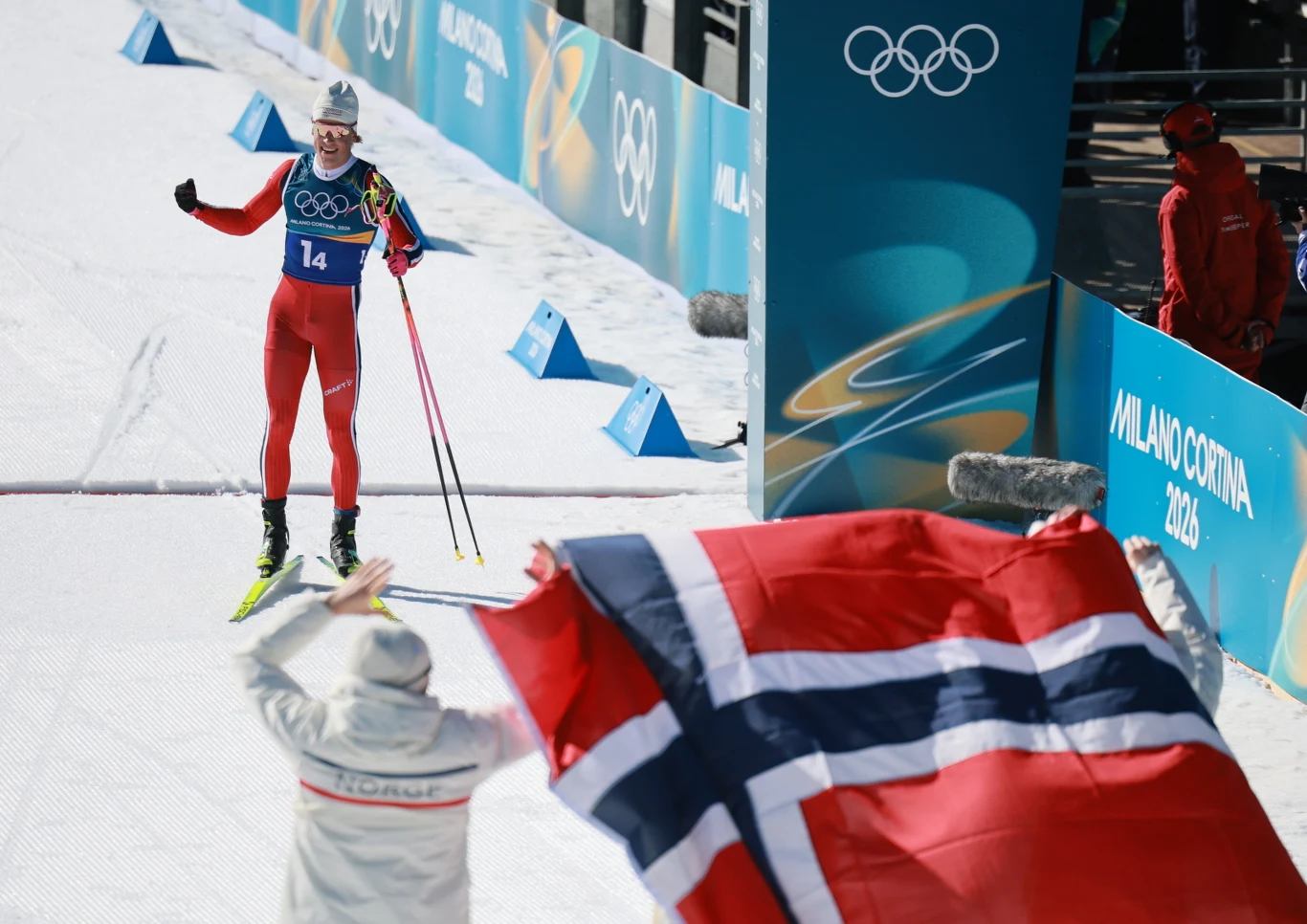 Narciarz biegowy w czerwonym stroju z numerem 14 finiszuje podczas zawodów olimpijskich, a w tle widoczna jest flaga Norwegii trzymana przez kibica oraz banery z logo igrzysk w Mediolanie i Cortina.