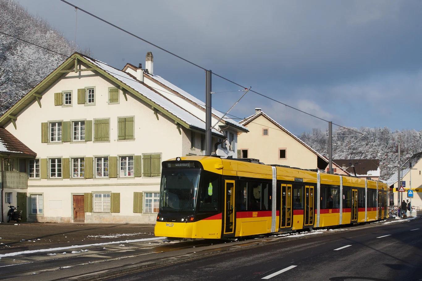 Szwajcaria uruchamiła pierwszą półautonomiczną kolej. Żółty nowoczesny tramwaj przejeżdża ulicą wśród zadbanych domów w stylu alpejskim, na tle lekko ośnieżonych drzew i nieba z chmurami.