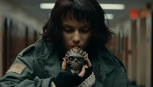 Młoda kobieta z krótkimi ciemnymi włosami trzyma w obu dłoniach dekoracyjną czaszkę i pochyla się, by ją pocałować, stojąc na tle pustego szkolnego korytarza z szafkami w tle. Ubrana jest w zieloną kurtkę i rękawiczki bez palców.
