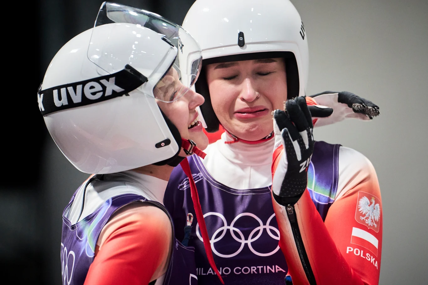 Dwie zawodniczki w kaskach narciarskich i kombinezonach olimpijskich świętują emocjonalny moment. Jedna z nich płacze ze wzruszenia, a druga okazuje jej wsparcie, obejmując ją z uśmiechem. Na stroju widoczne logo igrzysk olimpijskich oraz polska flaga.