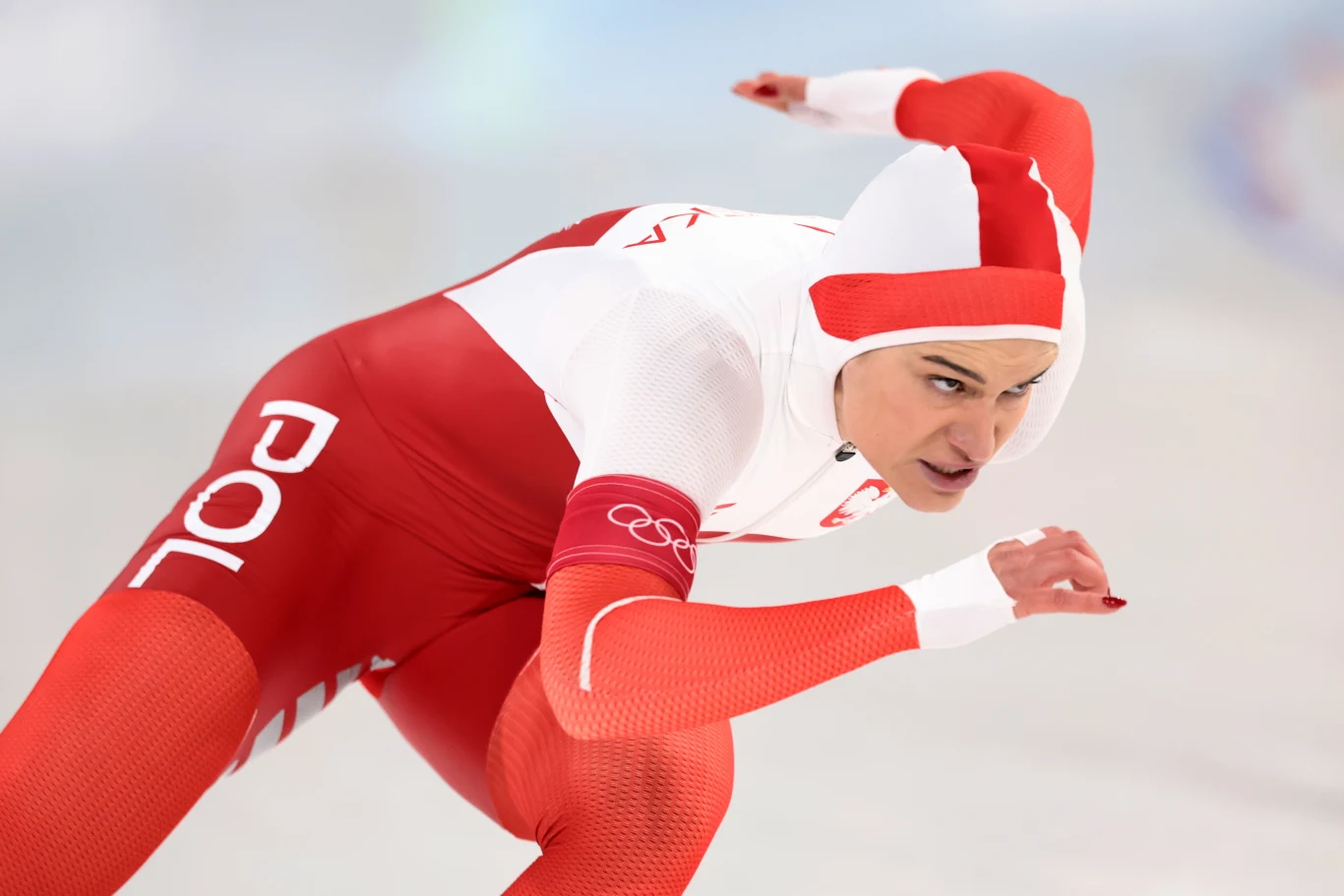 Andżelika Wójcik Zawodnik w biało-czerwonym stroju reprezentującym Polskę podczas wyścigu łyżwiarskiego, pochylony do przodu z wyciągniętą ręką, dynamiczny ruch na lodowej tafli, wyraz koncentracji na twarzy.