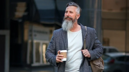 Mężczyzna z brodą, w płaszczu, z kubkiem kawy i plecakiem