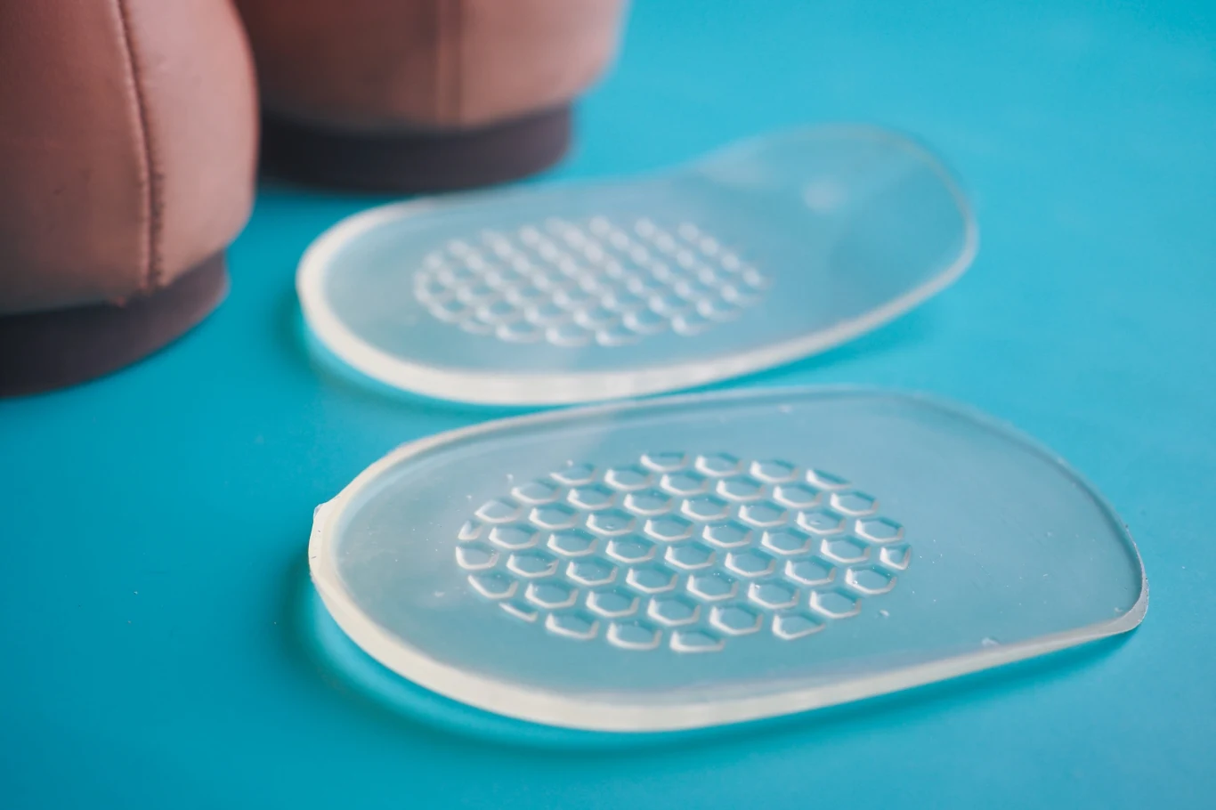 Podpiętki znajdziemy w formie żelowej lub skórzanej Dwie silikonowe wkładki do butów o przezroczystej strukturze z wypustkami ułożonymi w regularne wzory, na tle niebieskiej powierzchni, w tle fragment butów.