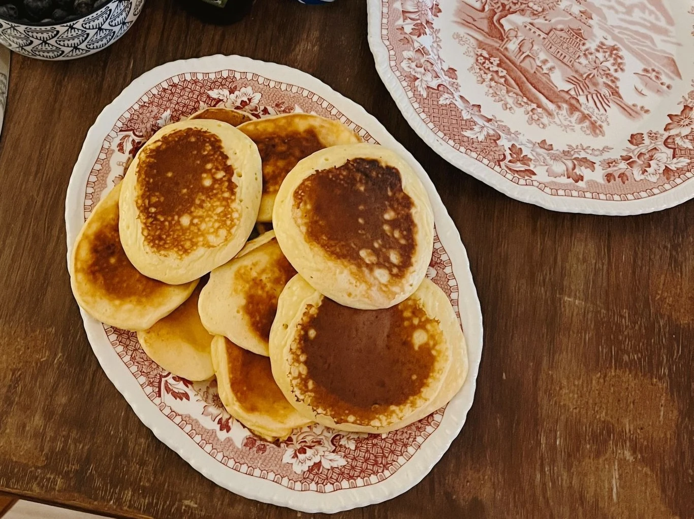 Stos puchatych, złocistych placuszków ułożony na ozdobnym półmisku na drewnianym stole, obok widoczny fragment talerza z dekoracyjnym wzorem.