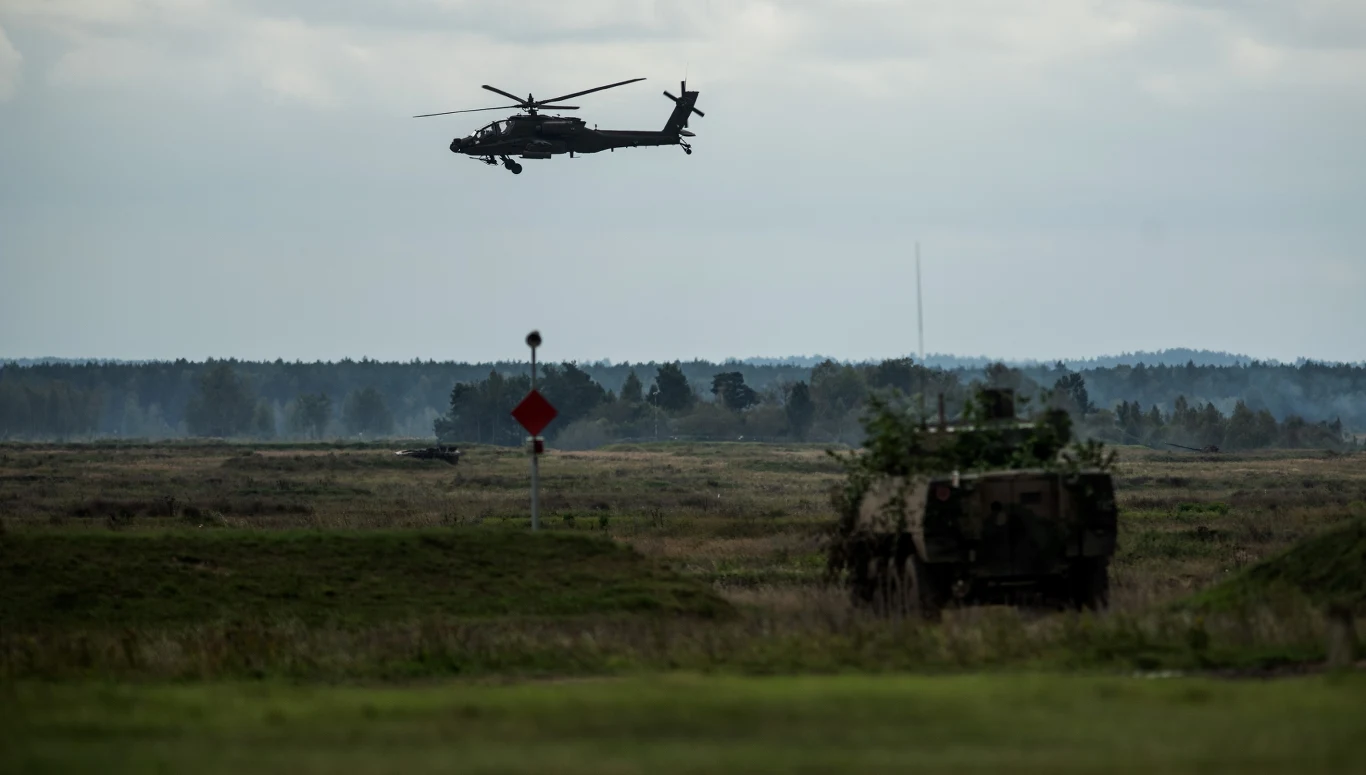 Program SAFE ma wesprzeć europejską obronność; zdj. ilustracyjne Śmigłowiec wojskowy unosi się nad poligonem, na pierwszym planie pojazd opancerzony częściowo zakamuflowany gałęziami, rozległa przestrzeń i zabudowania lasu w tle.