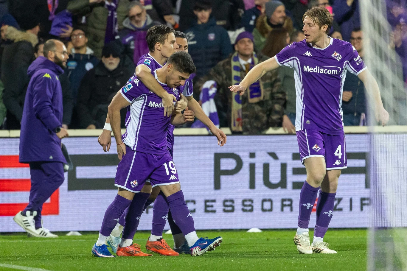 Zawodnicy drużyny piłkarskiej w fioletowych strojach celebrują wspólnie sukces na boisku, w tle widoczna publiczność oraz elementy stadionu.