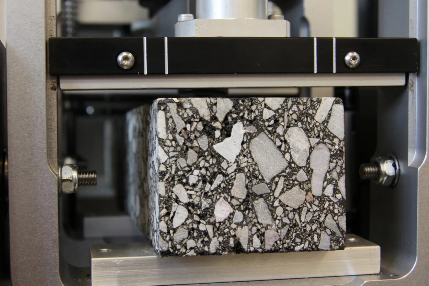 Bryła asfaltobetonu poddawana testowi w prasie laboratoryjnej. Widoczne ziarna kruszywa zatopione w masie bitumicznej, całość umieszczona w metalowej ramie urządzenia pomiarowego.