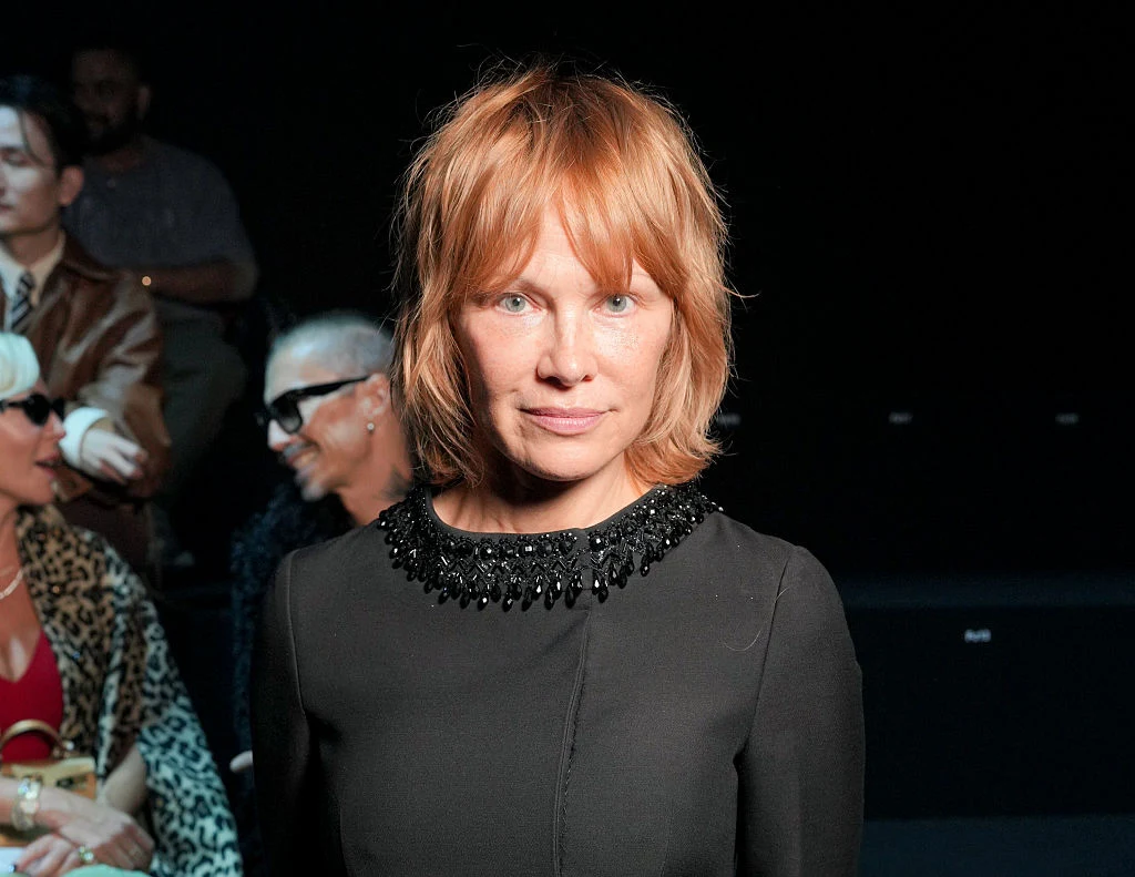 Pamela Anderson, 5 października 2025 roku podczas paryskiego tygodnia mody Kobieta o rudych włosach w czarnej sukience ozdobionej koralikami stoi na tle ciemnego wnętrza, w tle widoczne osoby siedzące na widowni.