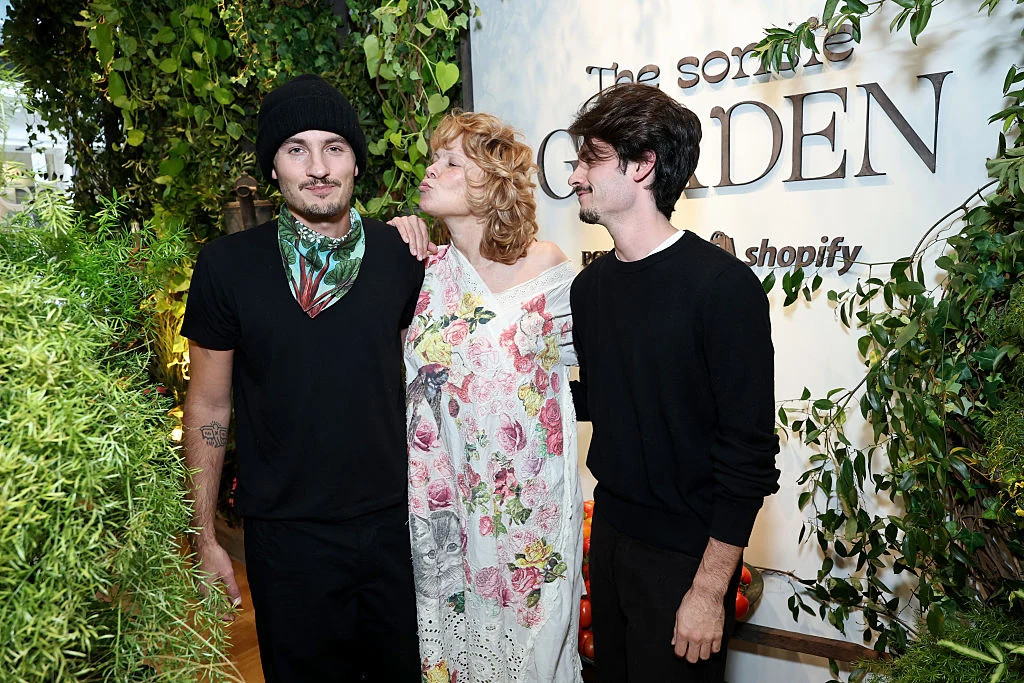 Pamela Anderson z synami Dylanem i Brandonem Lee podczas Sonsie Garden Trzech młodych dorosłych stoi razem w bujnej, zielonej scenerii, dwoje mężczyzn ubranych jest na czarno, a kobieta w jasnej, kwiecistej sukni uśmiecha się i trzyma ich za ramiona. W tle widoczny jest fragment napisu 'The some Garden' i logo Shopify.