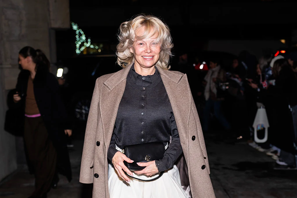 Pamela Anderson podczas nowojorskiego tygodnia mody Uśmiechnięta kobieta o jasnych włosach w eleganckim płaszczu i czarnej koszuli stoi na ulicy wieczorem. W tle grupa ludzi i oświetlone miejsca.