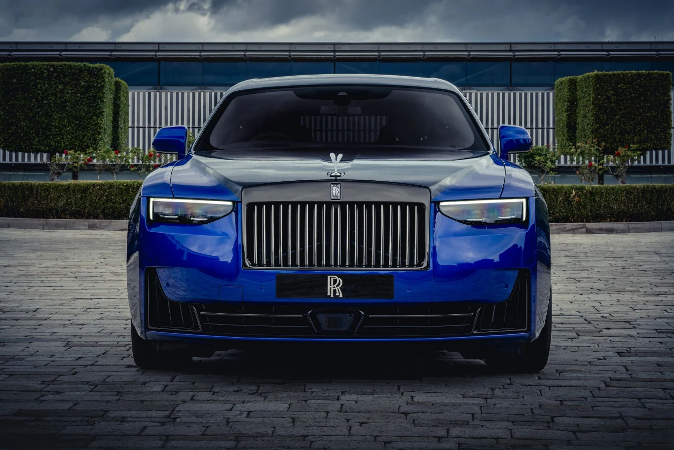 Luksusowy niebieski samochód marki Rolls-Royce zaparkowany na brukowanej alejce, widoczny z przodu, z wyraźnym chromowanym grillem oraz charakterystycznym emblematem na masce, w tle nowoczesny budynek i równo przystrzyżone żywopłoty.