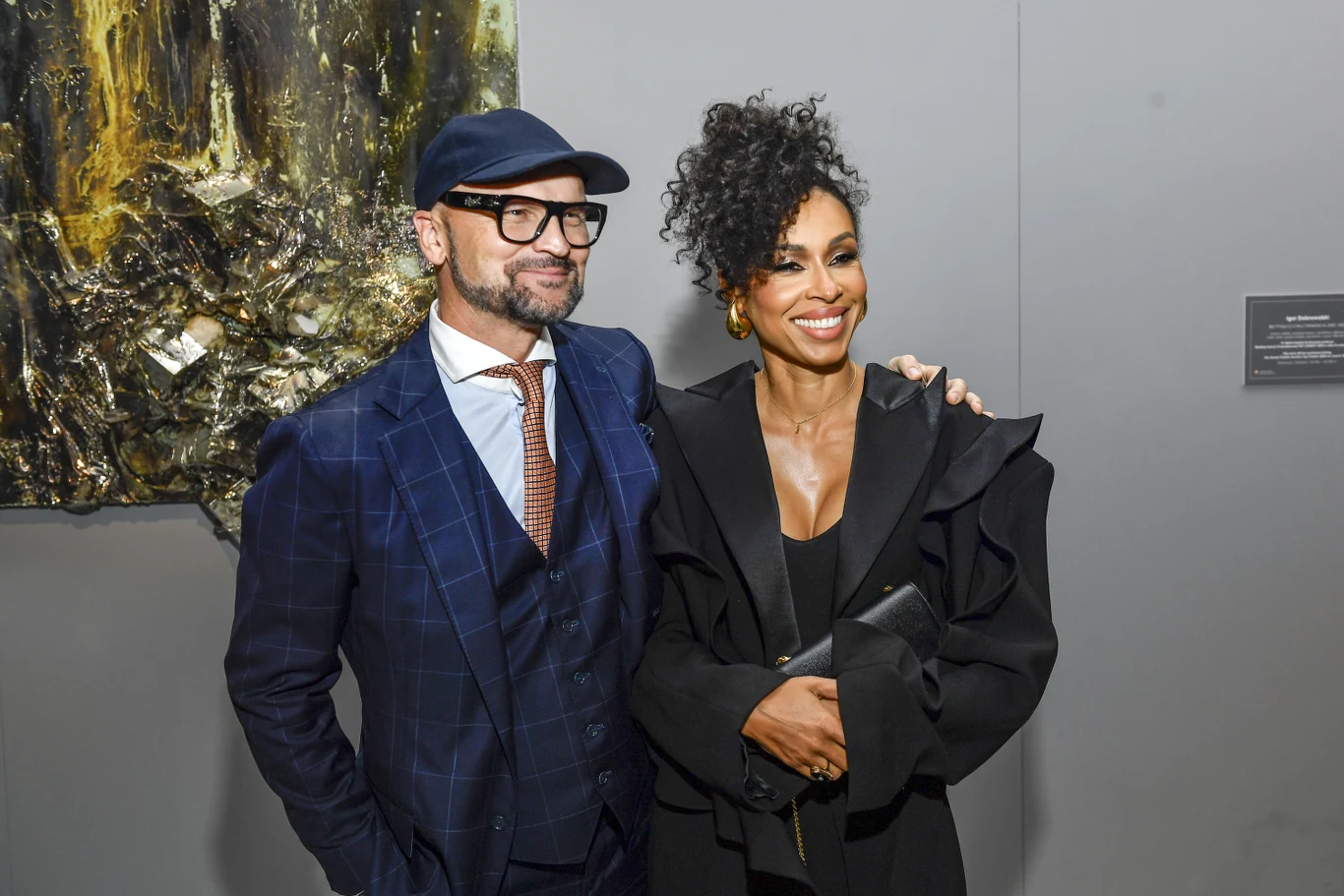 Dwie elegancko ubrane osoby pozujące razem na tle dzieła sztuki, mężczyzna w garniturze z okularami i czapką oraz kobieta w czarnej stylizacji z upiętymi włosami, oboje uśmiechnięci i zrelaksowani.