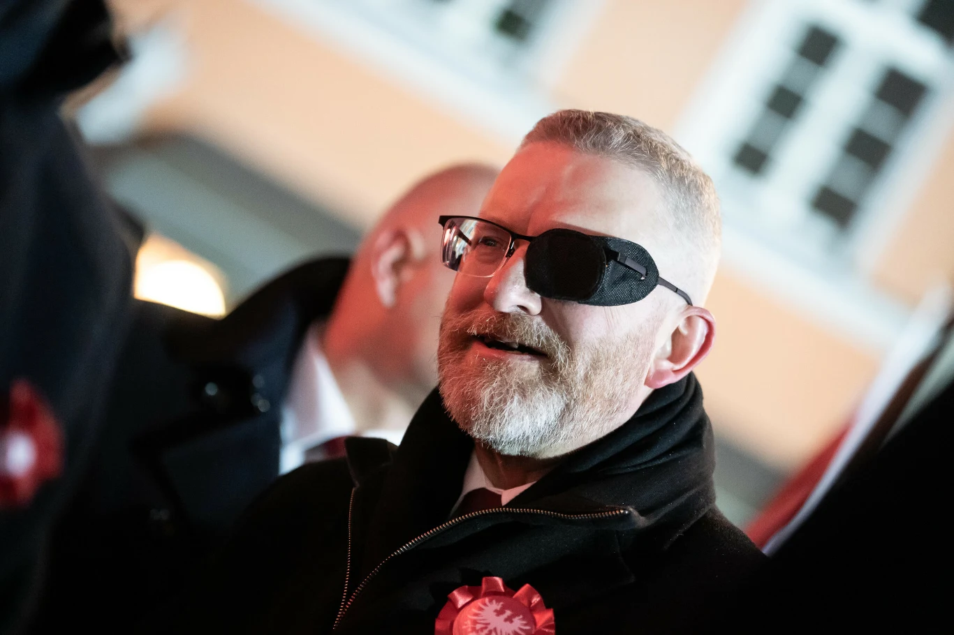 Mężczyzna z opaską na oku i brodą, ubrany w czarny płaszcz, na którym przypięta jest biało-czerwona kokarda, stojący na tle budynku z oknami.