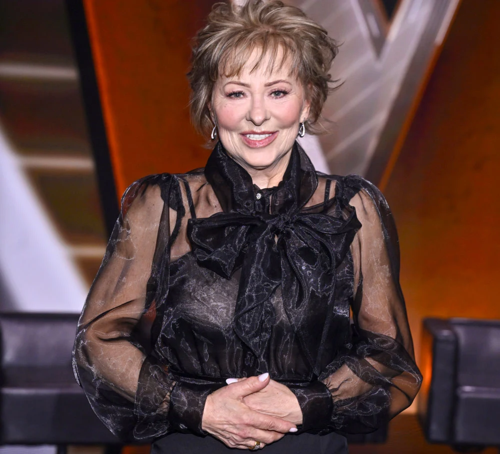 Grażyna Osmala dotarła do ścisłego finału "The Voice Senior" Kobieta w średnim wieku uśmiecha się stojąc na tle sceny, ubrana jest w elegancką, czarną bluzkę z dużą kokardą i przezroczystymi rękawami. Jej włosy są krótko ścięte i naturalnie ułożone.