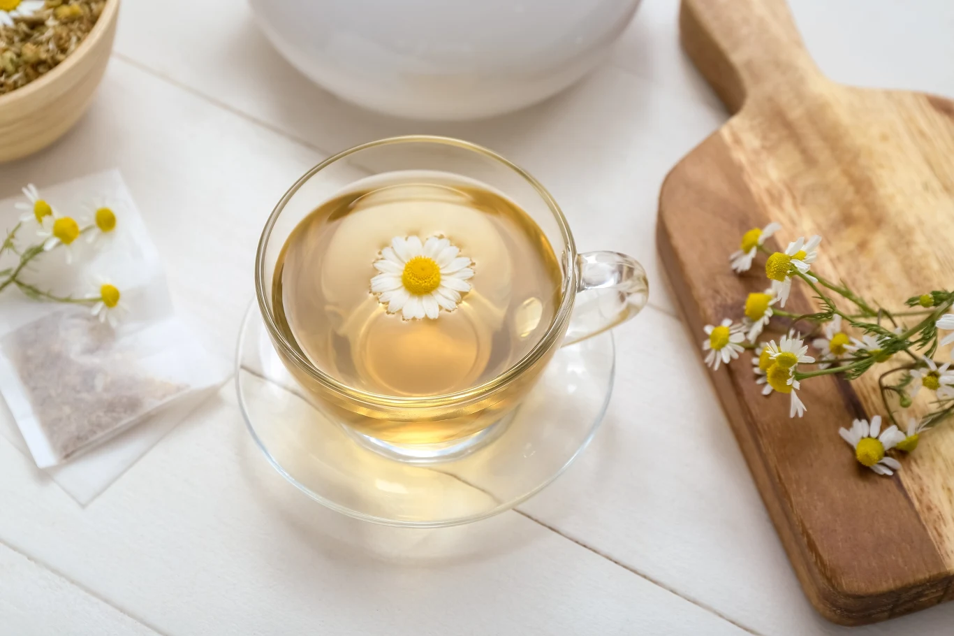 Szklanka z przejrzystym naparem z rumianku, na jej powierzchni unosi się kwiat rumianku. Obok znajduje się drewniana deska z kilkoma kwiatami oraz saszetka herbaty i miseczka z suszonym rumiankiem.