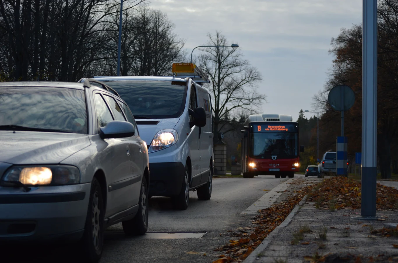 Szara osobówka oraz biały van poruszają się ulicą obok krawężnika, w oddali widoczny nadjeżdżający autobus oraz jesienne drzewa bez liści, część jezdni pokryta suchymi liśćmi.