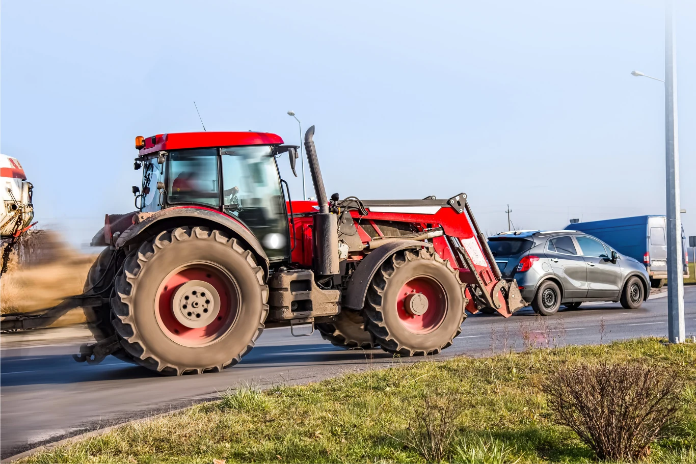 Czerwony ciągnik rolniczy poruszający się po asfaltowej drodze obok srebrnego samochodu osobowego, w tle widać elementy zabudowań oraz krzewy na pierwszym planie.