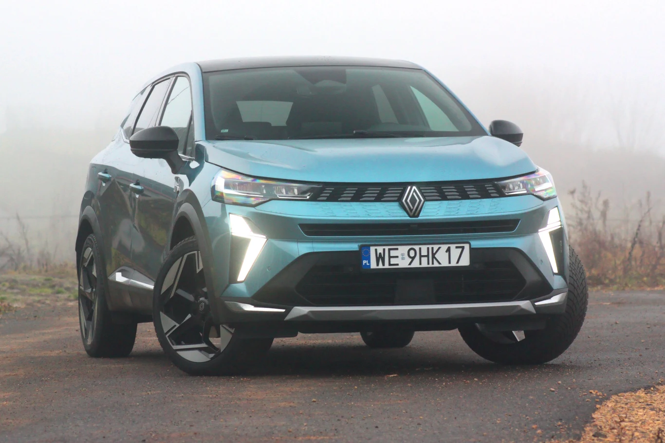 Renault Symbioz to model, który zastąpił Megane Nowoczesny samochód osobowy koloru niebiesko-szarego z wyraźnymi światłami LED, widoczny od frontu na tle mgły, stojący na asfaltowej nawierzchni. Na pojeździe widoczna tablica rejestracyjna z polskim numerem.