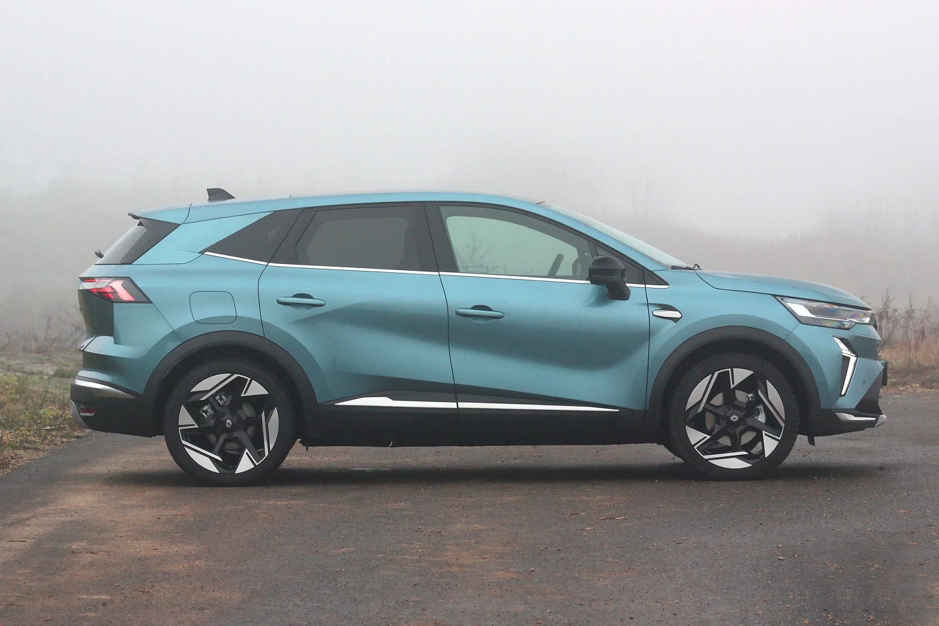 Renault Symbioz to naprawdę ładny, dynamicznie wyglądający samochód Niebieski SUV nowoczesnej stylistyki stoi zaparkowany na asfaltowej drodze w otoczeniu lekkiej mgły, widoczny z profilu, koła aluminiowe i sportowe elementy nadwozia podkreślają dynamiczny wygląd pojazdu.