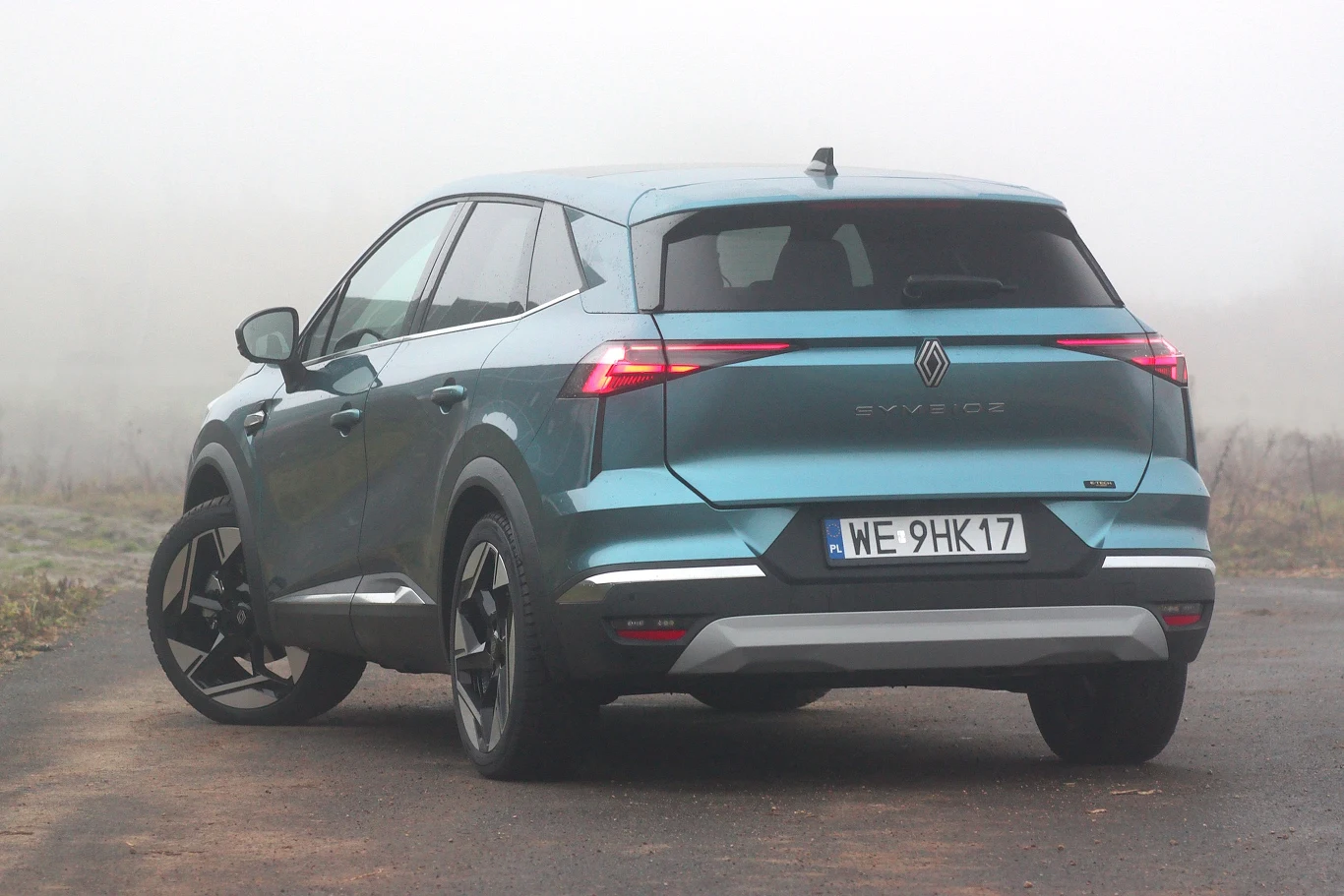 Renault Symbioz Nowoczesny, niebieski SUV marki Renault Austral stoi na asfaltowej drodze w otoczeniu gęstej mgły. Samochód widoczny jest od tyłu, z wyróżniającymi się światłami LED, czarnymi felgami oraz tablicą rejestracyjną WE 9HK17.