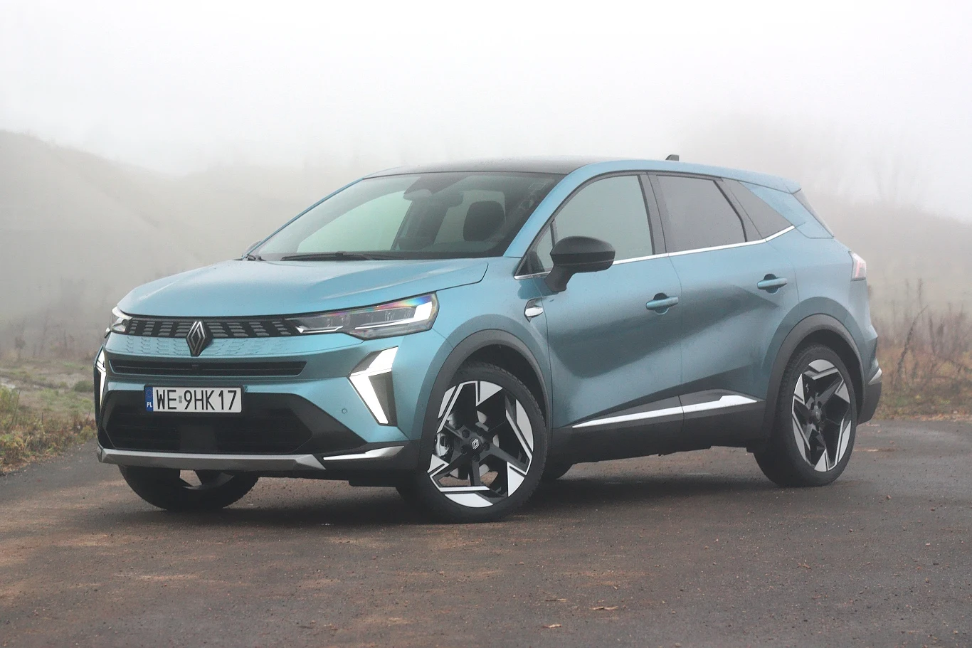 Nowoczesny, niebieski samochód typu SUV stoi na asfaltowej drodze, w tle mglista sceneria oraz niewyraźne zarysy roślinności i wzgórz. Auto wyróżnia się wyrazistymi felgami i charakterystycznym designem świateł przednich.