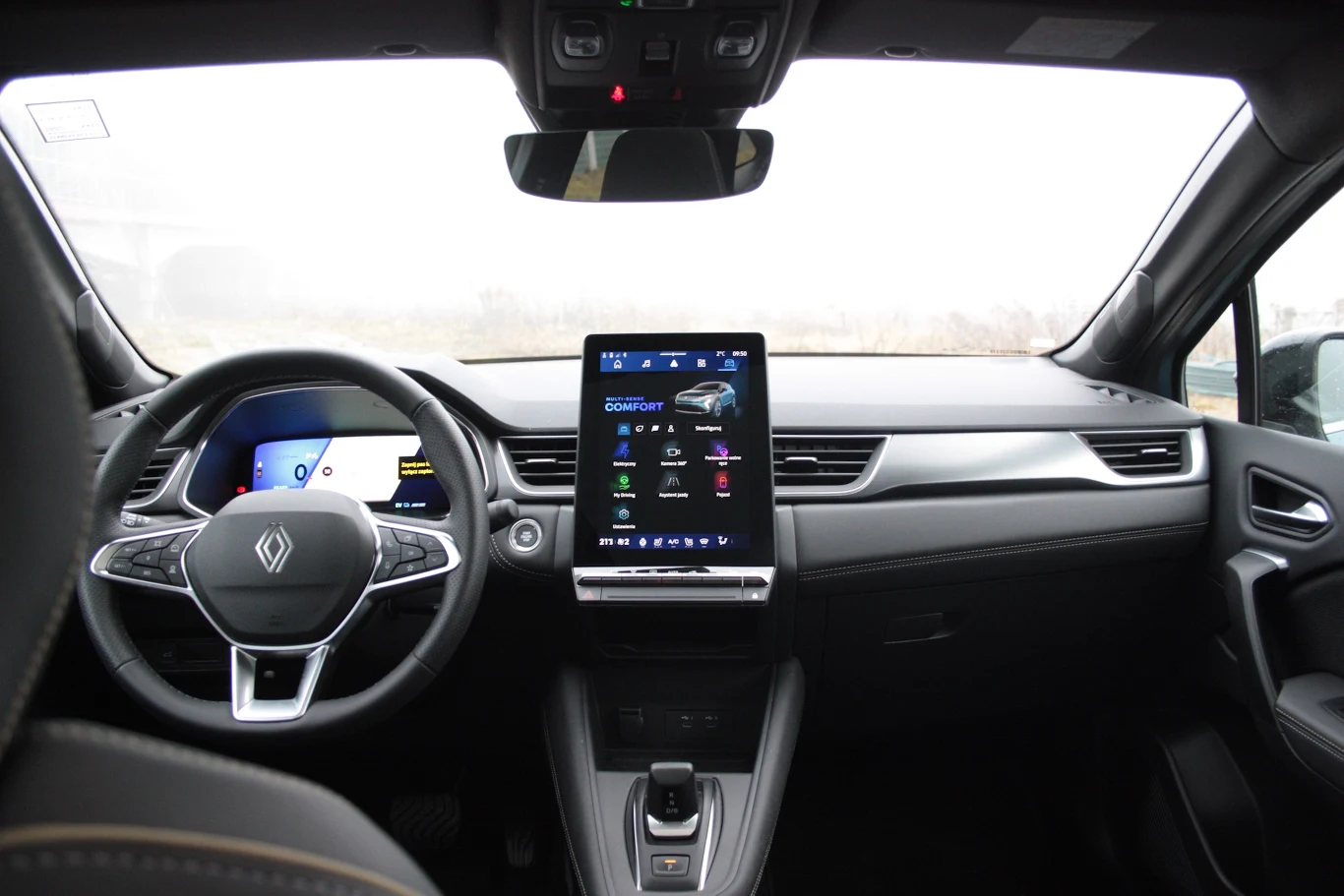 Kabina Renault Symbioz jest zdominowana przez centralny wolnostojący ekran Nowoczesna deska rozdzielcza samochodu Renault z cyfrowym wyświetlaczem za kierownicą oraz dużym ekranem dotykowym w centralnej części kokpitu. Wnętrze utrzymane w ciemnych kolorach, z widocznym logo marki na kierownicy.