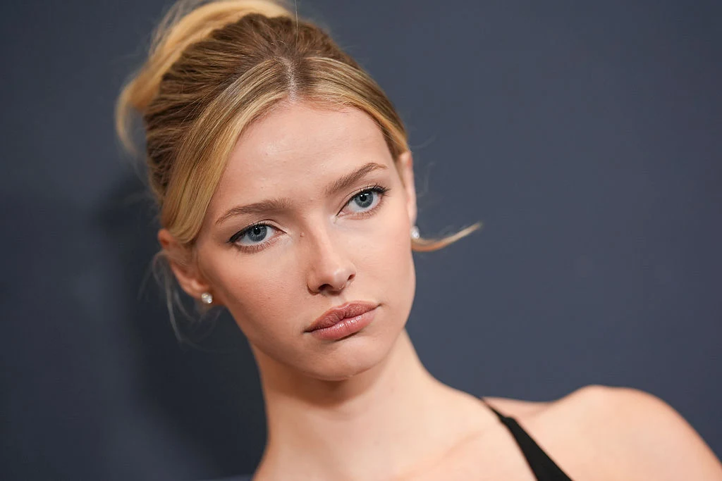 Apple Martin Młoda kobieta o jasnych włosach spiętych w kok, o jasnej cerze, delikatnym makijażu i niebieskich oczach, patrząca z lekkim zamyśleniem na ciemnym, jednolitym tle.