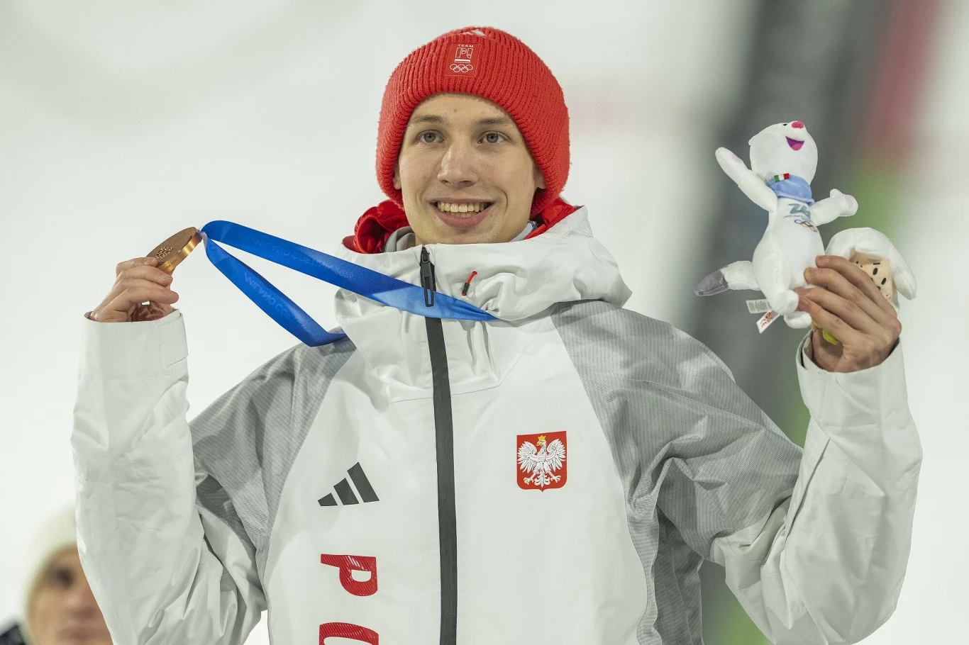 Kacper Tomasiak Sportowiec w zimowej kurtce z orzełkiem Polski na piersi trzyma medal oraz pluszową maskotkę, uśmiecha się i nosi czerwoną czapkę.