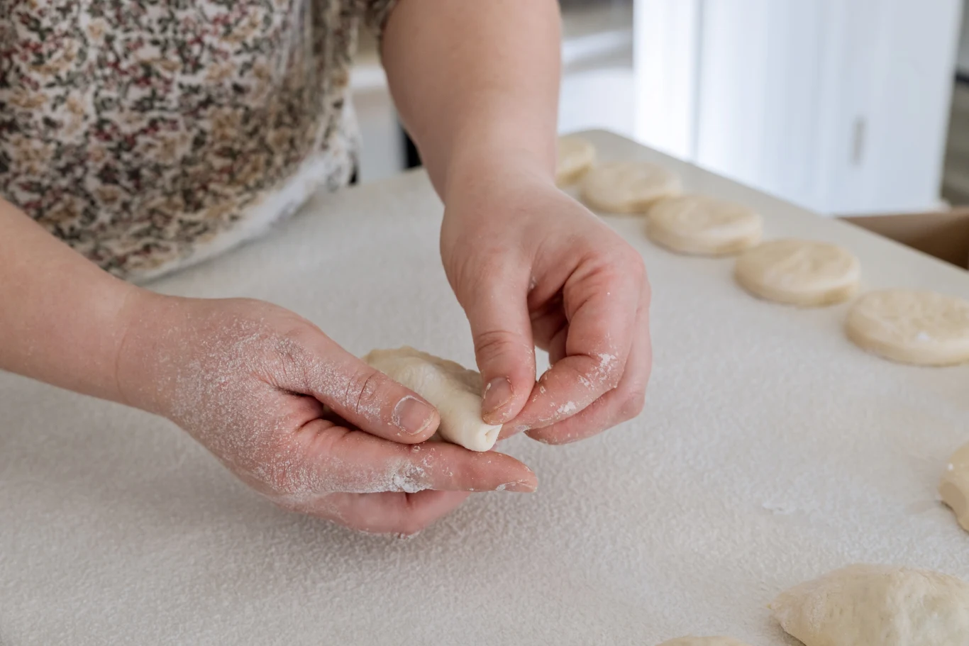 Ręce osoby formujące ciasto na jasnej powierzchni, w tle leżą równo ułożone kawałki surowego ciasta.