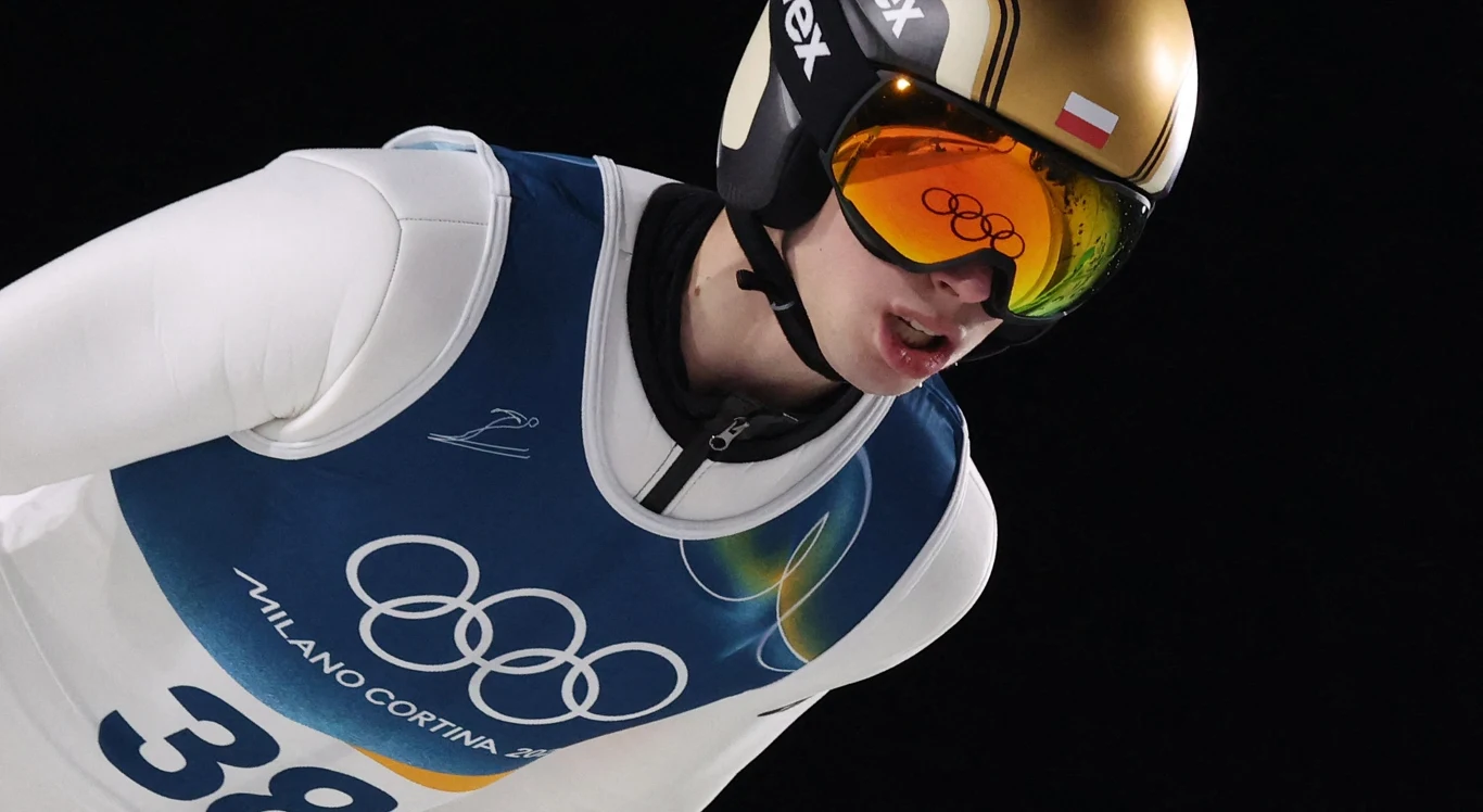 Skoczek narciarski w stroju startowym z numerem 38, kaskiem i goglami z logo olimpijskim, pochylony do przodu podczas skoku na tle czerni, wyraźnie widoczne logo igrzysk olimpijskich oraz polska flaga na kasku.