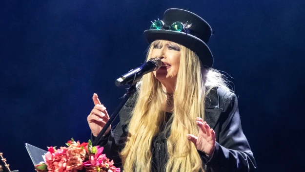 Kobieta w ozdobionym kapeluszu i efektownym stroju śpiewa na scenie trzymając mikrofon, w tle instrumenty muzyczne i kwiatowa dekoracja przy statywie.