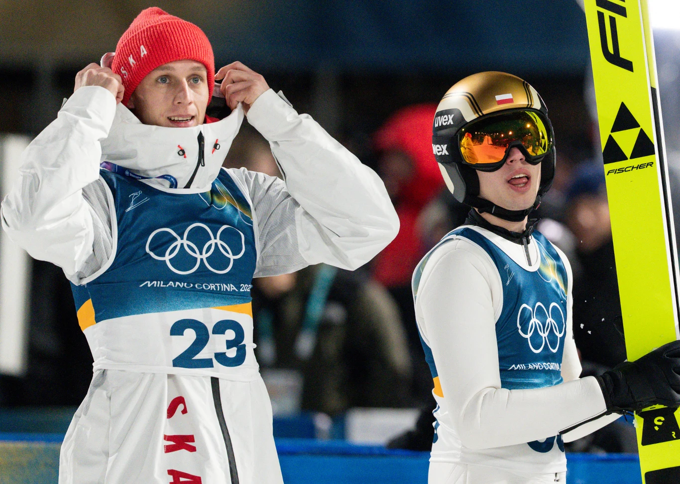 Skoki narciarskie. Na zdjęciu Paweł Wąsek i Kacper Tomasiak Dwóch skoczków narciarskich w kombinezonach sportowych, jeden w czerwonej czapce poprawia kurtkę z widocznym numerem startowym 23, drugi trzyma narty w jednej ręce, obaj mają gogle ochronne i zimowe ubrania z logo igrzysk olimpijskich.