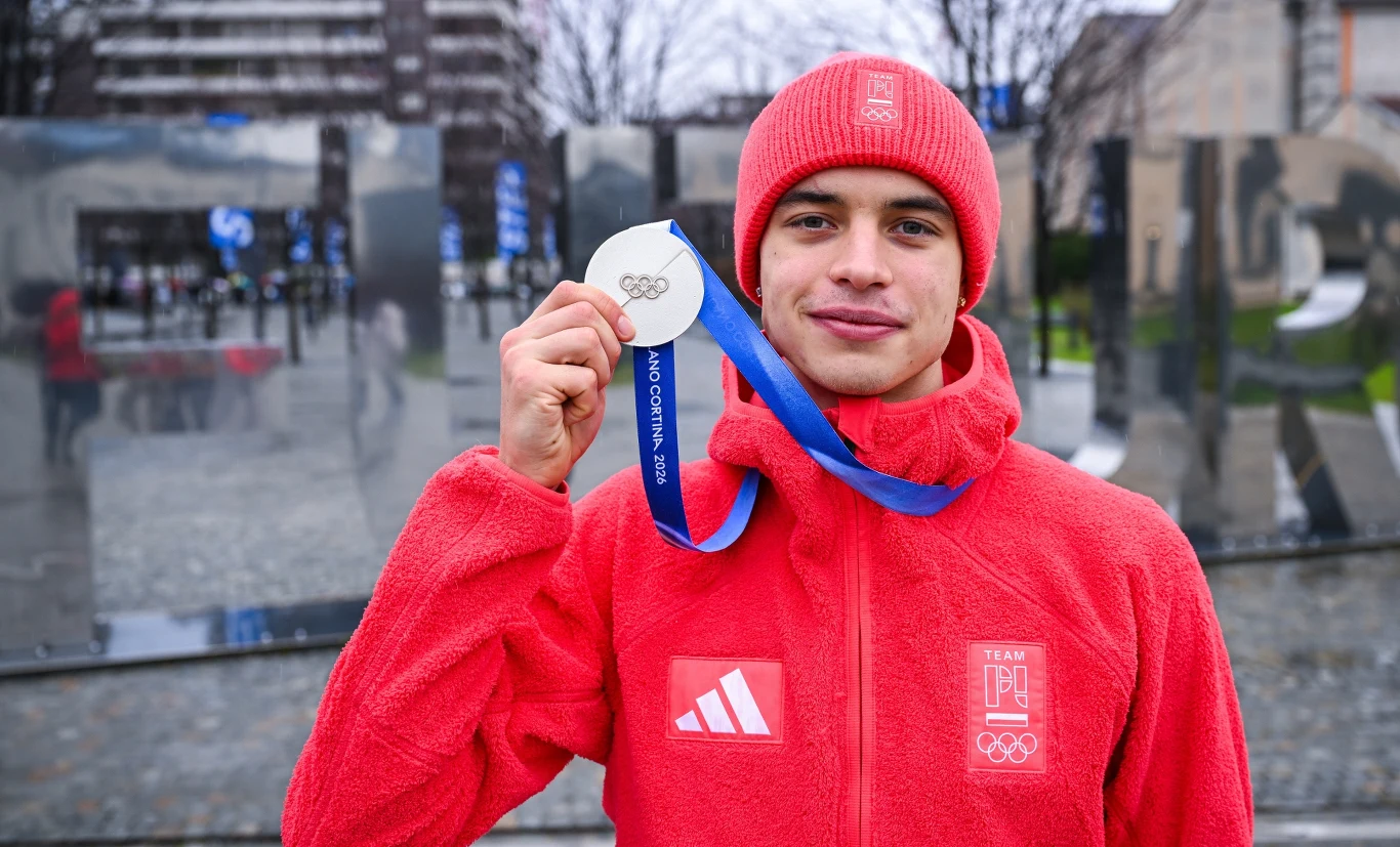 Władimir Siemirunnij Młody sportowiec w czerwonym stroju trzyma w dłoni srebrny medal olimpijski i uśmiecha się do kamery, w tle nowoczesne budynki i szklana instalacja.