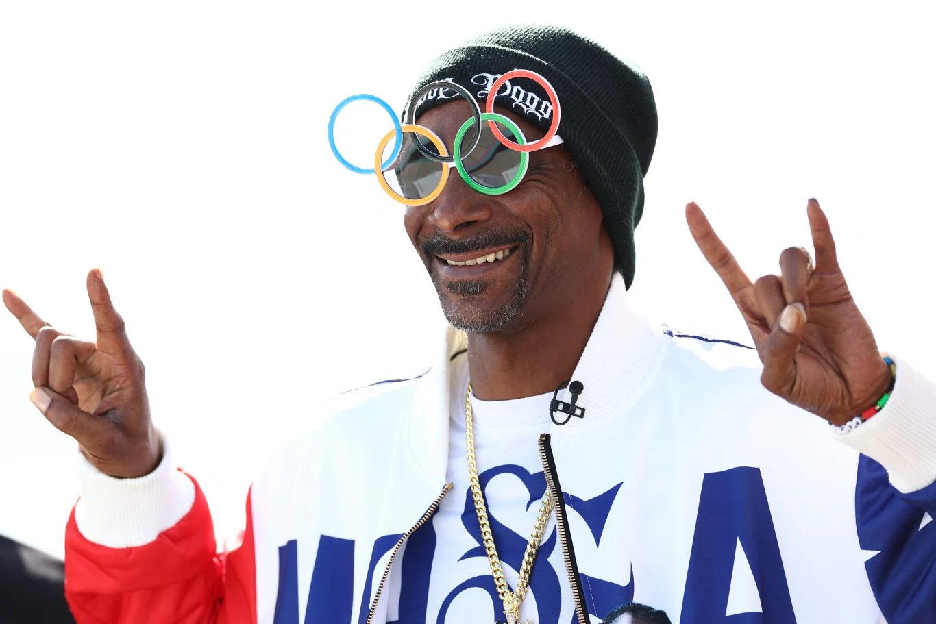 Snoop Dogg Uśmiechnięty mężczyzna w czapce i charakterystycznych okularach w kształcie kół olimpijskich pozuje z podniesionymi dłońmi, układając palce w znak "rock on". Ubrany w biało-niebiesko-czerwoną kurtkę z widocznym napisem, na szyi złoty łańcuch.