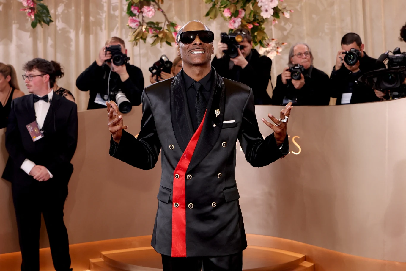 Snoop Dogg Elegancko ubrany mężczyzna w czarnej marynarce z czerwonym akcentem i okularach przeciwsłonecznych pozuje do zdjęcia na tle jasnej, ozdobionej kwiatami ścianki, otoczony przez fotografów i innych gości uroczystości.