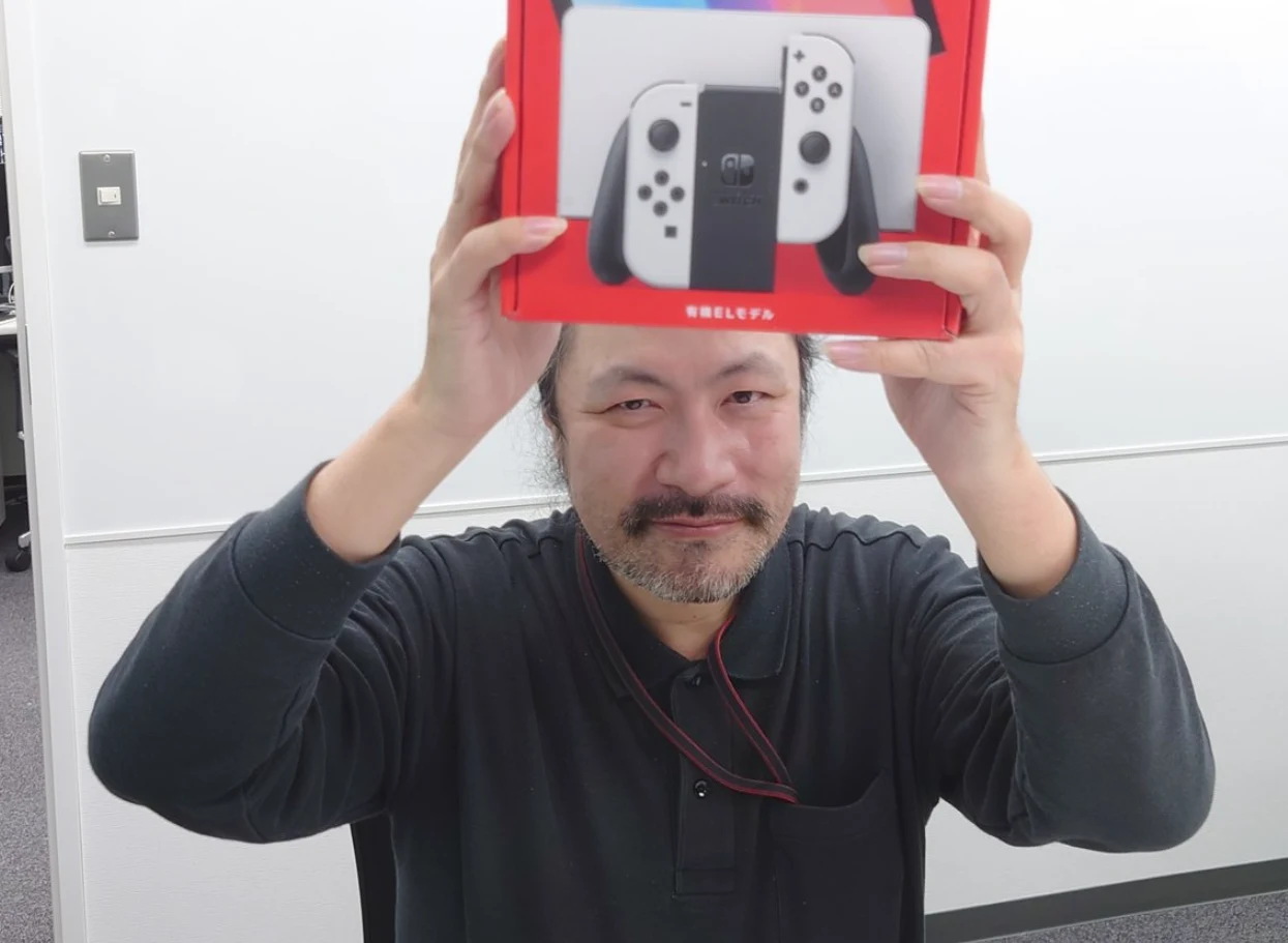 Shutao Iida Mężczyzna z zarostem trzyma nad głową czerwone pudełko z konsolą Nintendo Switch, uśmiechając się lekko. W tle widoczne są jasne ściany biura.