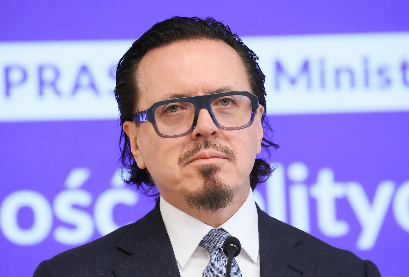 Ministerstwo Aktywów Państwowych, któremu szefuje Wojciech Balczun odpowiedziało na zarzuty Jacka Sasina. Mężczyzna w eleganckim garniturze i krawacie, z niebieskimi okularami, stoi przed mikrofonem na tle niebieskiej tablicy z białym napisem. Włosy zaczesane do tyłu, wyraz skupienia na twarzy.