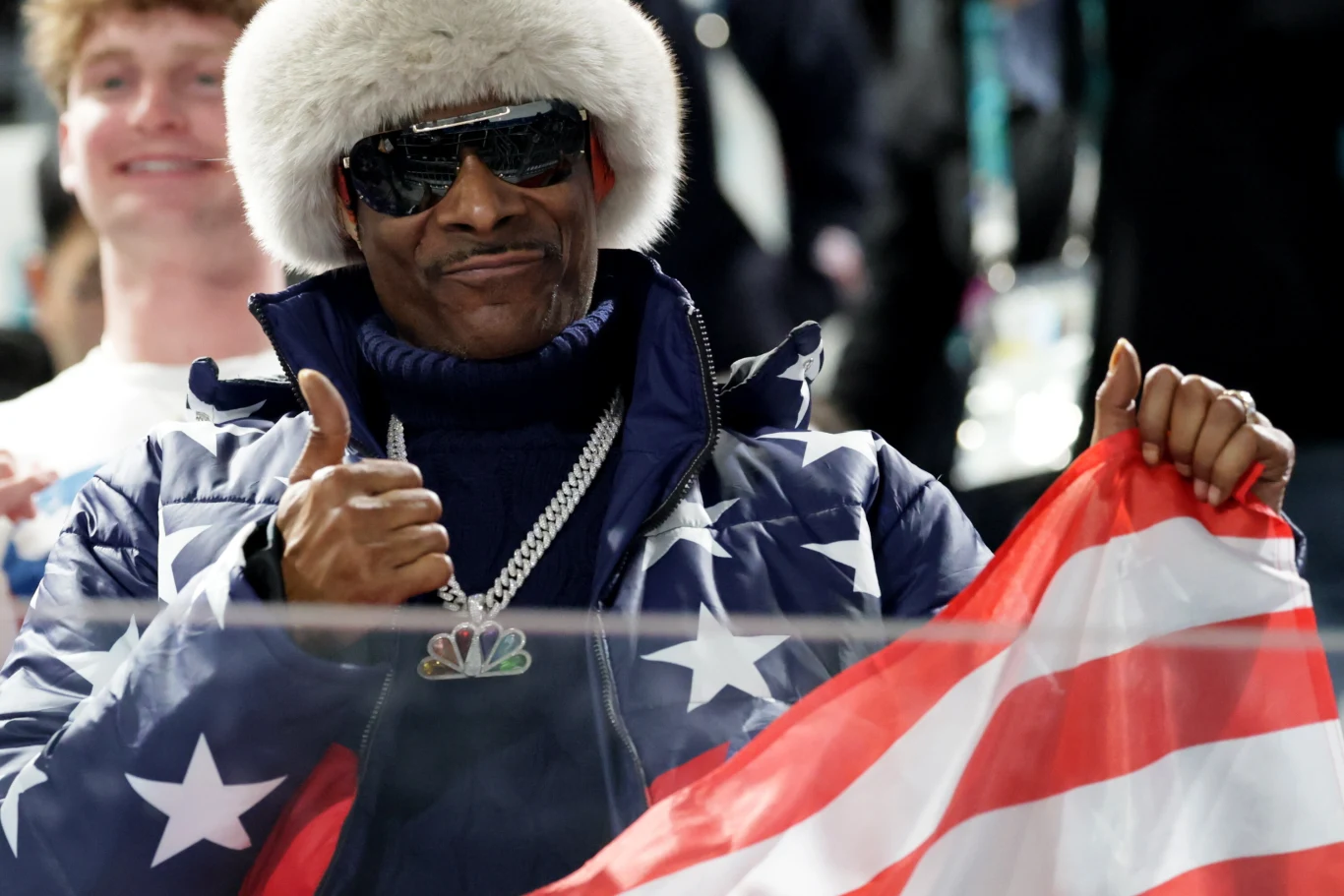 Snoop Dogg Mężczyzna w futrzanej czapce, okularach przeciwsłonecznych oraz kurtce w amerykańskie gwiazdy, trzymający flagę USA i pokazujący kciuk w górę.