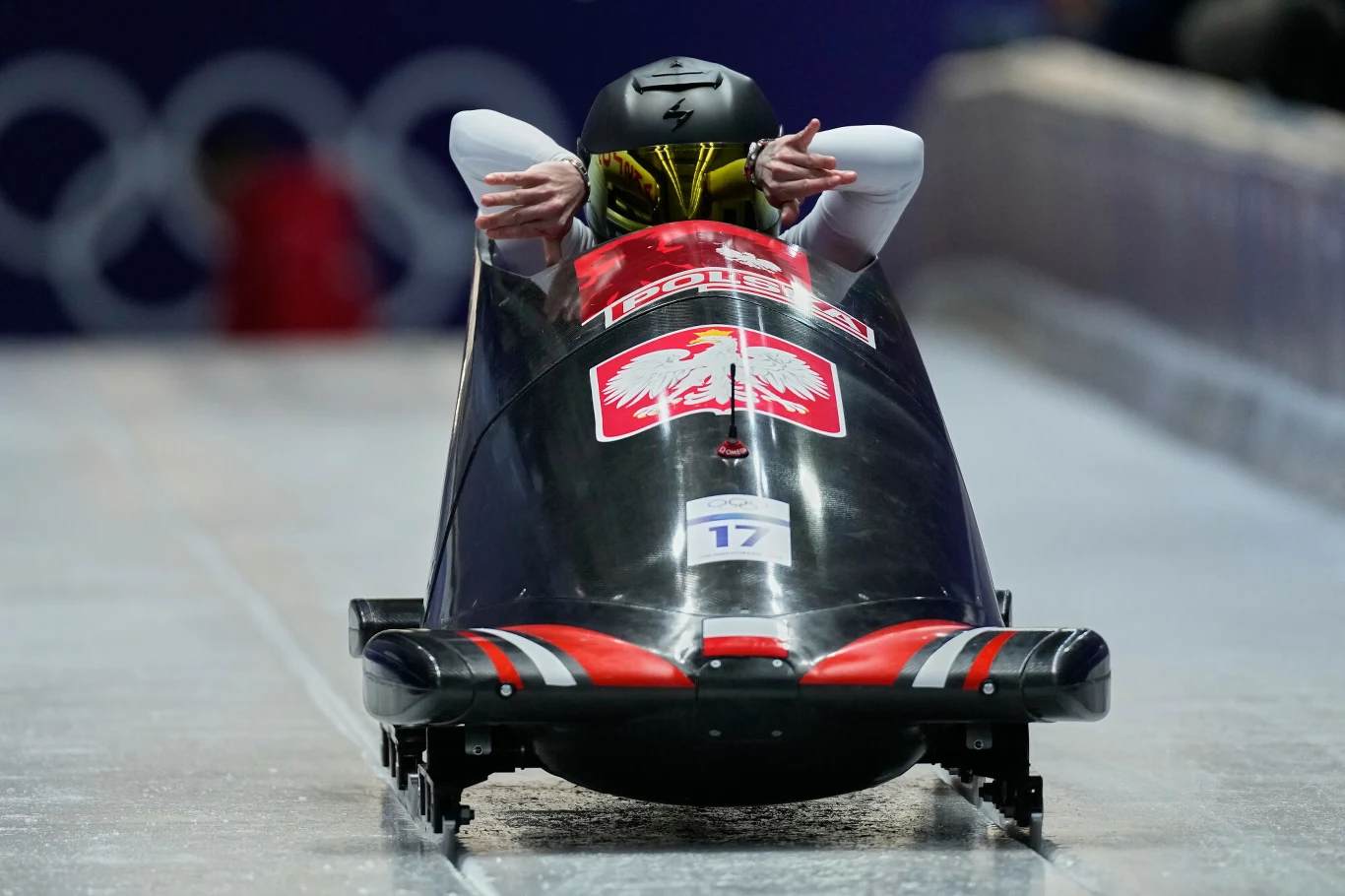 Linda Weiszewski Czteroosobowy polski bobslej na torze lodowym w trakcie wyścigu, na przodzie widoczna wyraźna flaga Polski, zawodnicy ubrani w sportowe kombinezony z kaskami.