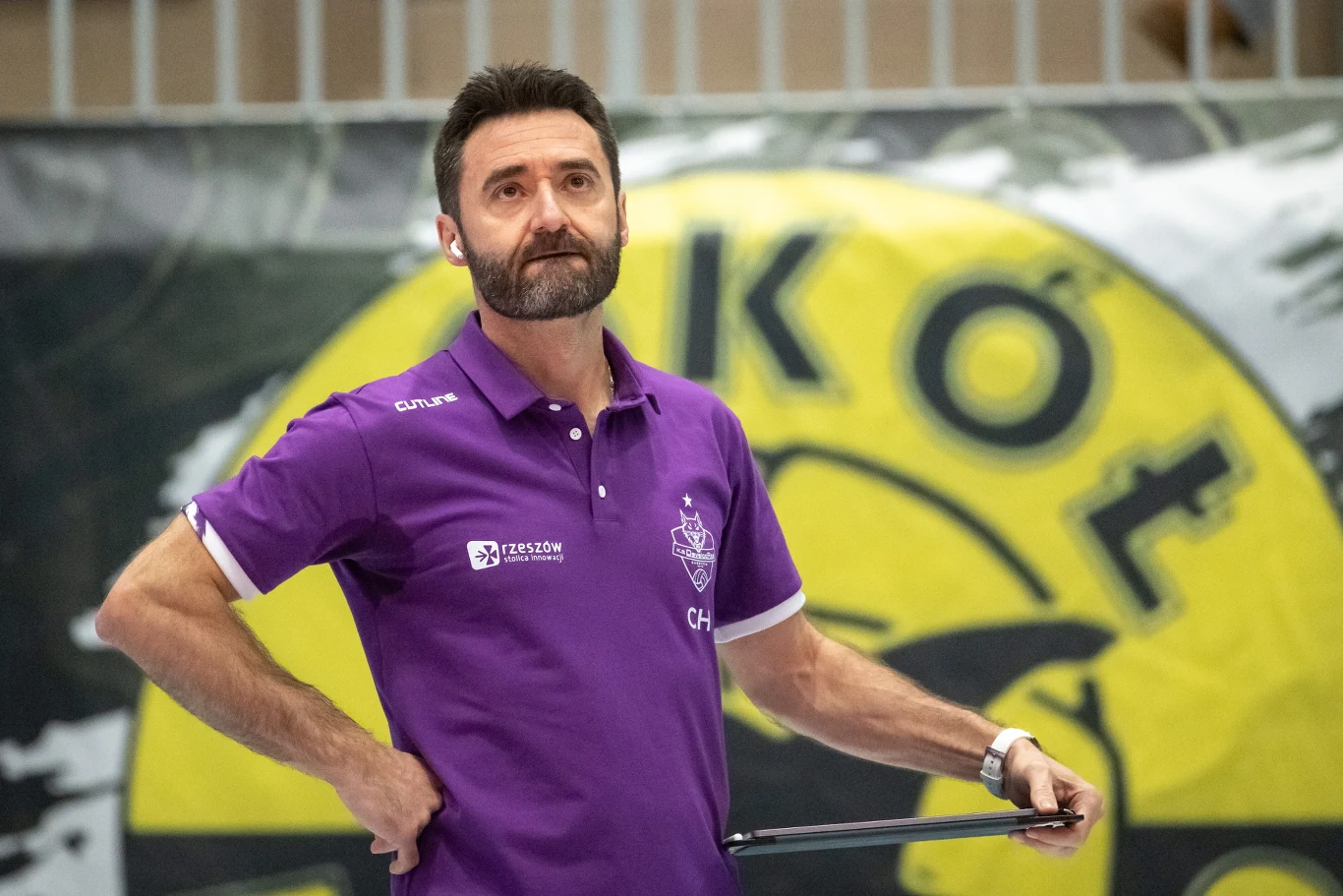 Mężczyzna z zarostem w fioletowej koszulce polo z logo drużyny sportowej stoi przed dużym żółto-czarnym banerem z napisem, trzymając coś w prawej dłoni.