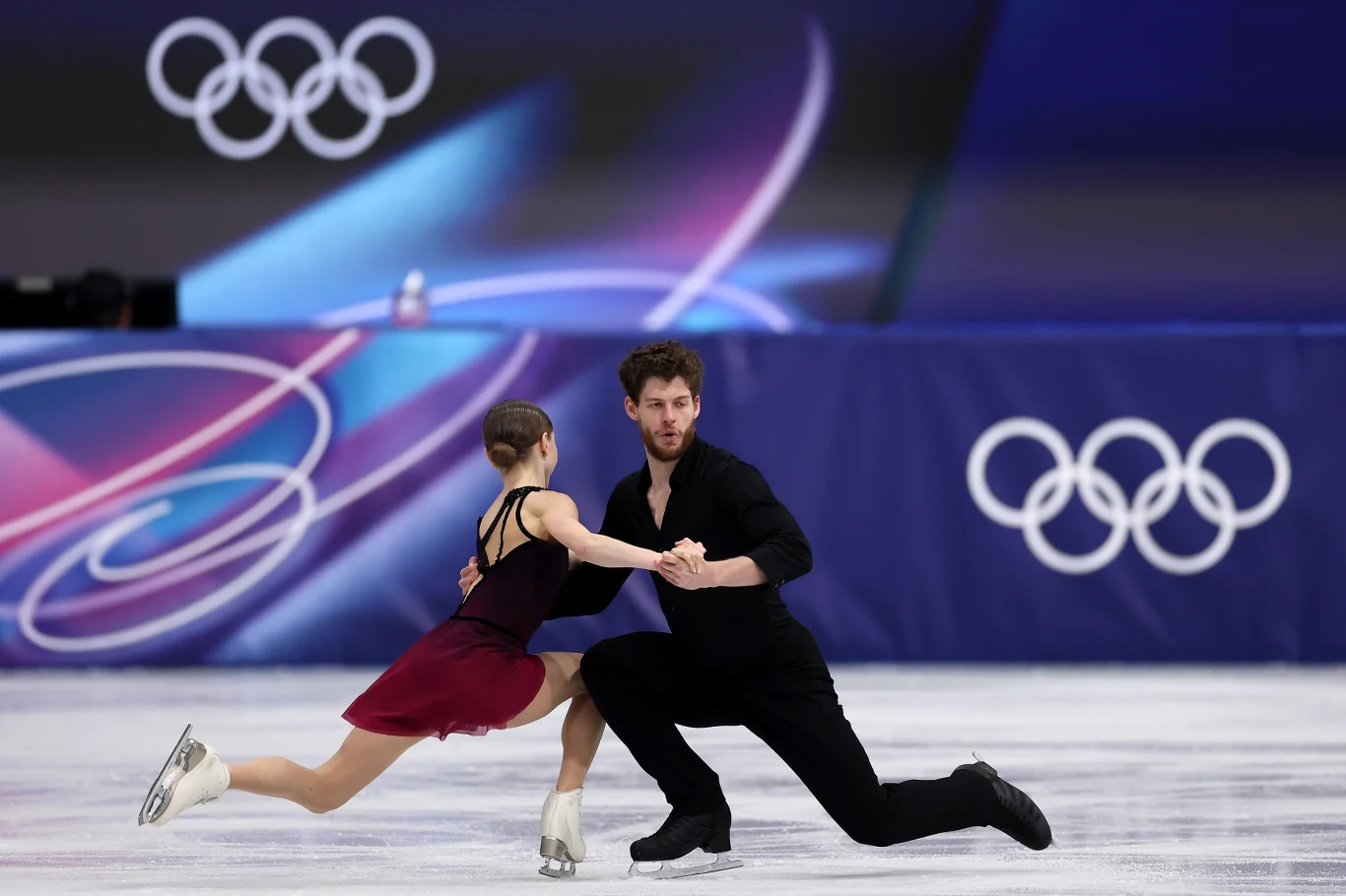 Julia Szczecinina i Michał Woźniak Para łyżwiarzy figurowych wykonuje wspólną figurę na lodowisku podczas zawodów olimpijskich, tło dekorują charakterystyczne symbole igrzysk.