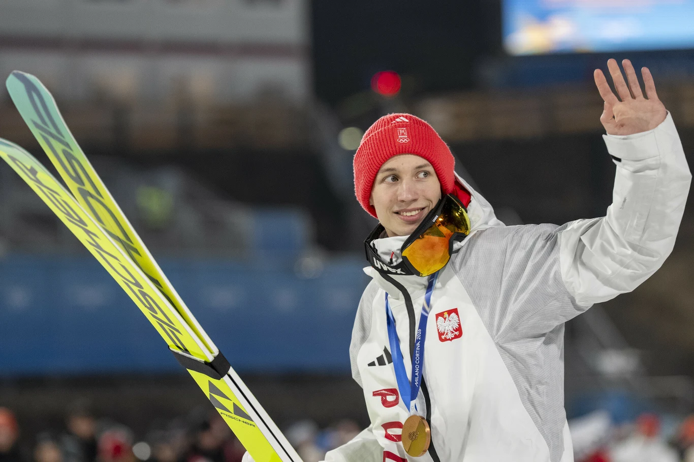 Sportowiec w białym kombinezonie i czerwonej czapce, z medalem na szyi oraz nartami w ręce, uśmiecha się i macha do publiczności podczas zawodów narciarskich. Na kurtce widoczne godło Polski.