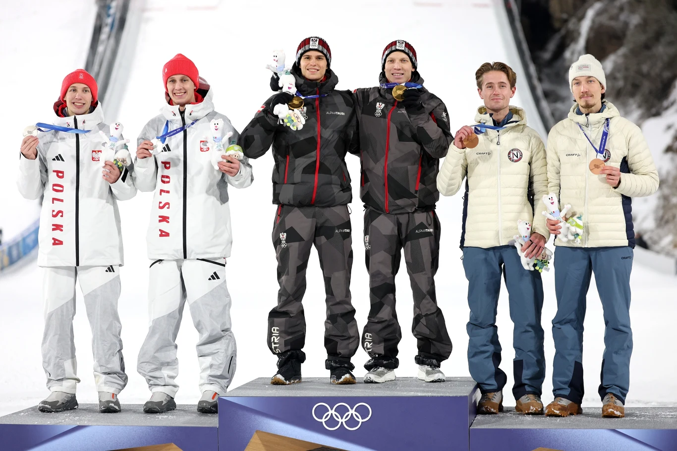 Medaliści zawodów olimpijskich w skokach narciarskich stoją na podium, trzymając medale i maskotki, w tle zeskok skoczni pokryty śniegiem.