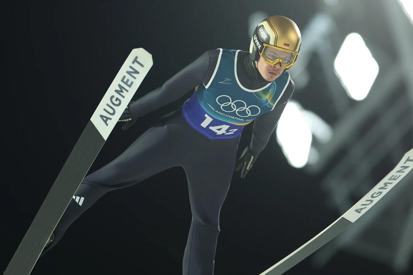 Skoczek narciarski w locie podczas zawodów olimpijskich, ubrany w ciemny kombinezon, złoty kask i gogle, widoczne narty oznaczone napisem 'AUGMENT', za zawodnikiem oświetlone tło nocą.