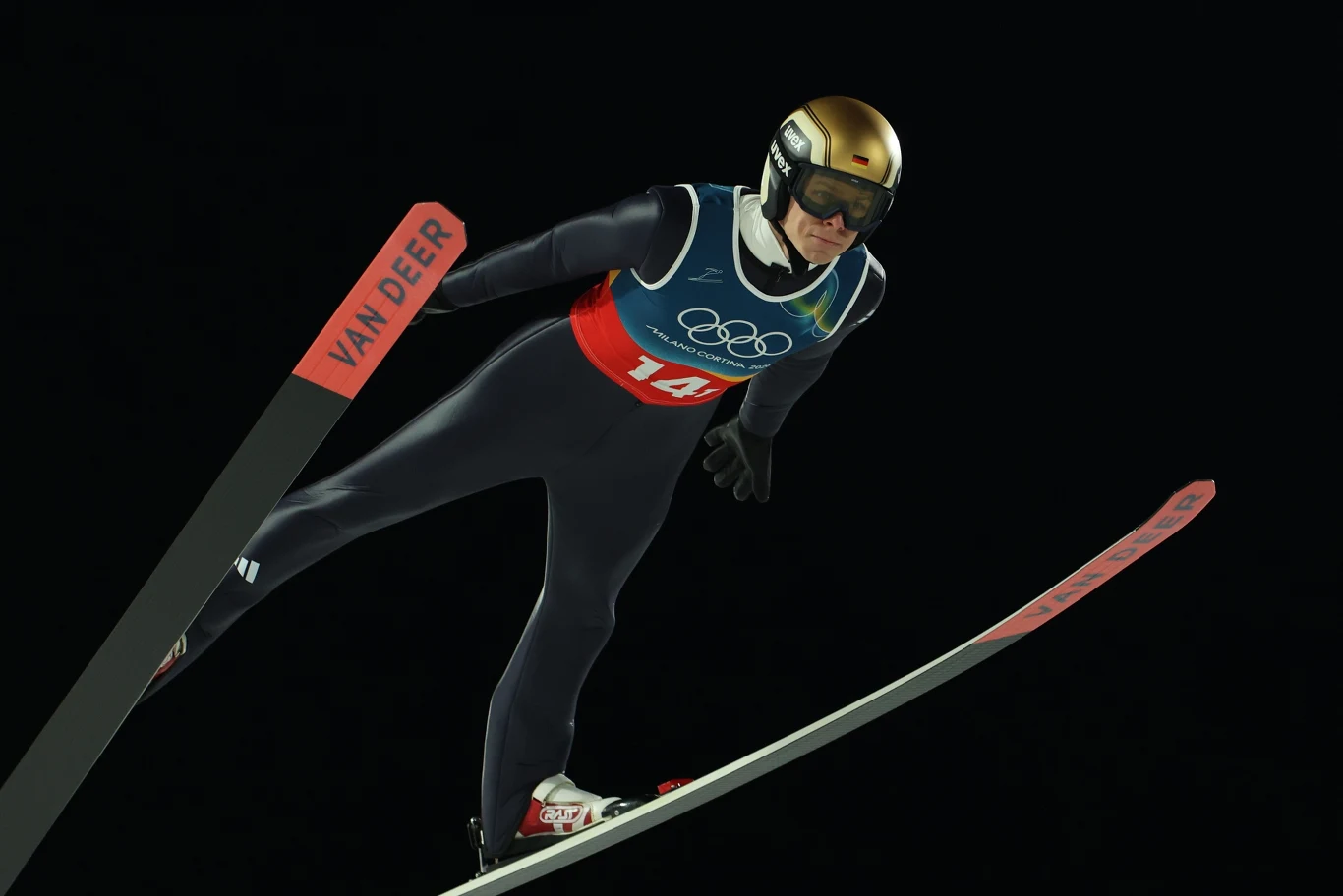 Skoczek narciarski wzbijający się w powietrze podczas konkursu olimpijskiego, ubran w kombinezon sportowy, kask i gogle, ciemne tło podkreśla dynamikę ruchu.