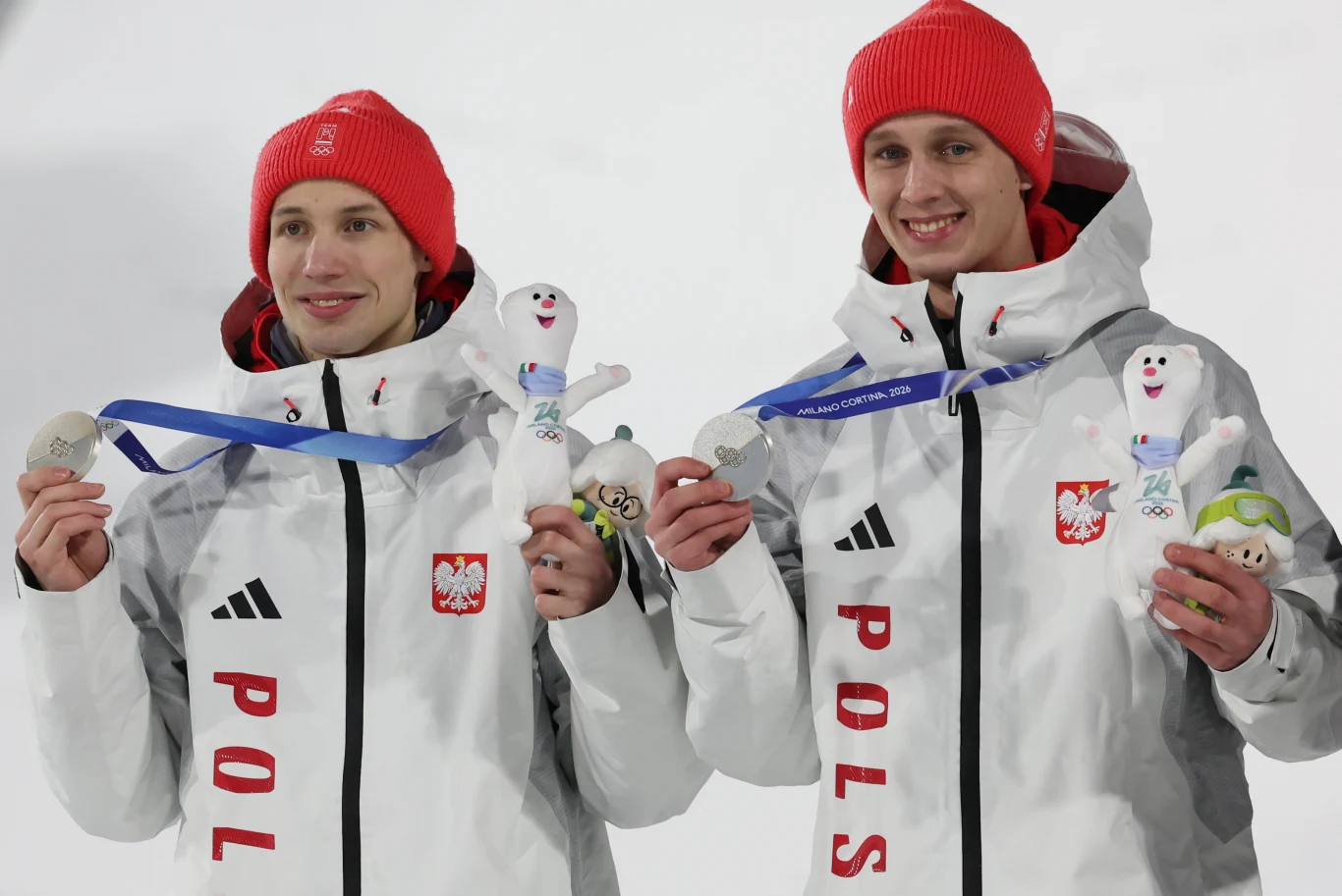 Dwóch sportowców w biało-czerwonych kurtkach z emblematem Polski i czerwonych czapkach trzyma srebrne medale oraz maskotki olimpijskie, obaj uśmiechnięci po zdobyciu nagród.