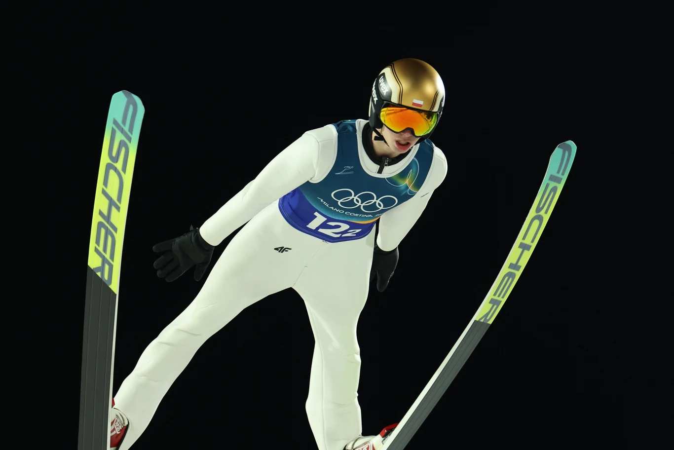 Skoczek narciarski w locie, ubrany w biały kombinezon i złoty kask, na tle ciemnego nieba, z widocznymi nartami marki Fischer oraz numerem startowym na piersi.